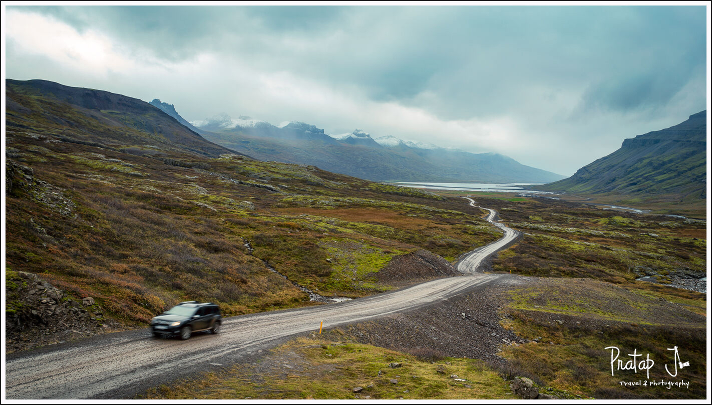 A winding road with a car on it and distant snow clad mountains in the East Fjords of Iceland